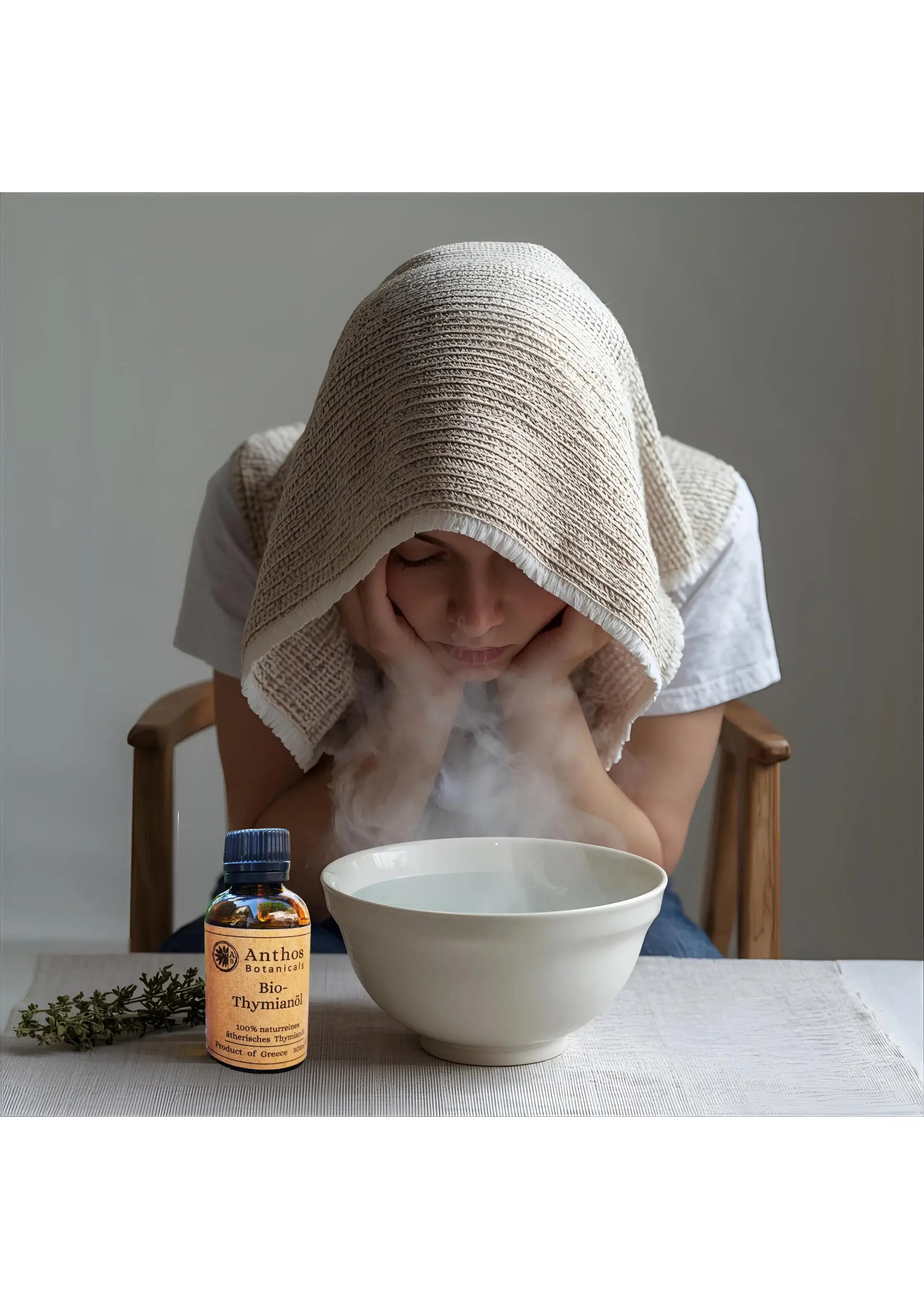 Eine junge Frau sitzt auf einem Stuhl mit einem Handtuch über dem Kopf und inhaliert ätherisches Thymianöl bio von Anthos Botanicals.
