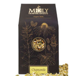 Eine schwarze Verpackung des Kamillenblütentee BIO. Ein Sichtfenster ist vorhanden und man erkennt Kamillenblüten. Kamillenblüten liegen vor und neben der Verpackung.