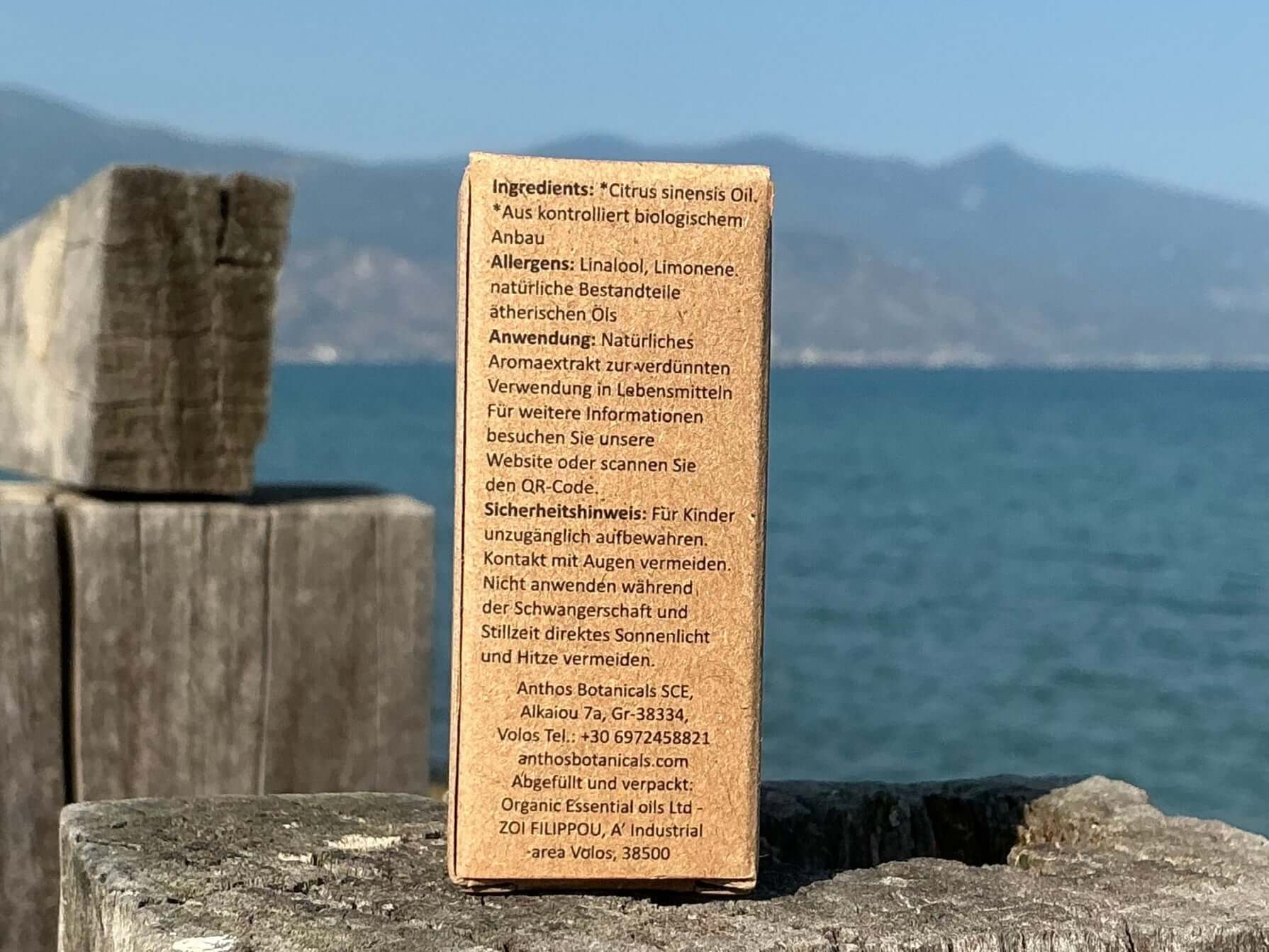 Rückseite einer braunen Kraftverpackung mit ätherischem Orangenöl Bio auf einem Holzsteg am Meer. Im Hintergrund das Peliongebirge.