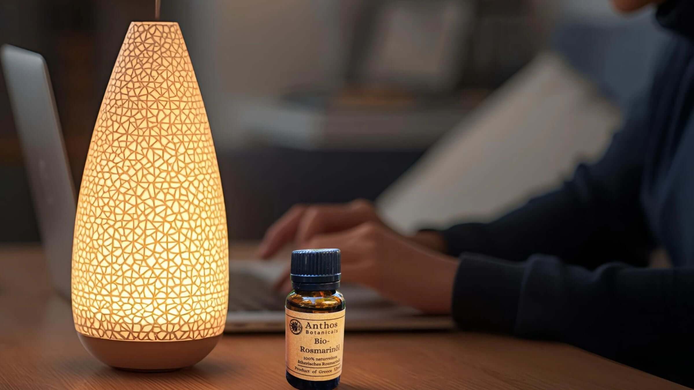 herobild eines Fläschens mit ätherischem Rosmarinöl vor einem Diffusor in einer Homeoffice-Szenerie