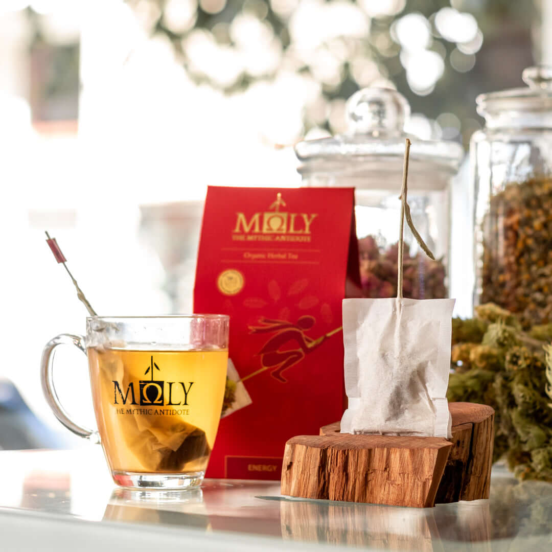 Eine rote Verpackung des Moly Energy Bio vor einem Hintergrund mit Vorratsgläsern an Kräutern. Zentral ist ein Bild aufgedruckt, welches einen Teebeutel mit Inhalt zeigt aus dem der Stengel der Sideritis scardia (griechischer Bergtee) herausragt und auf d