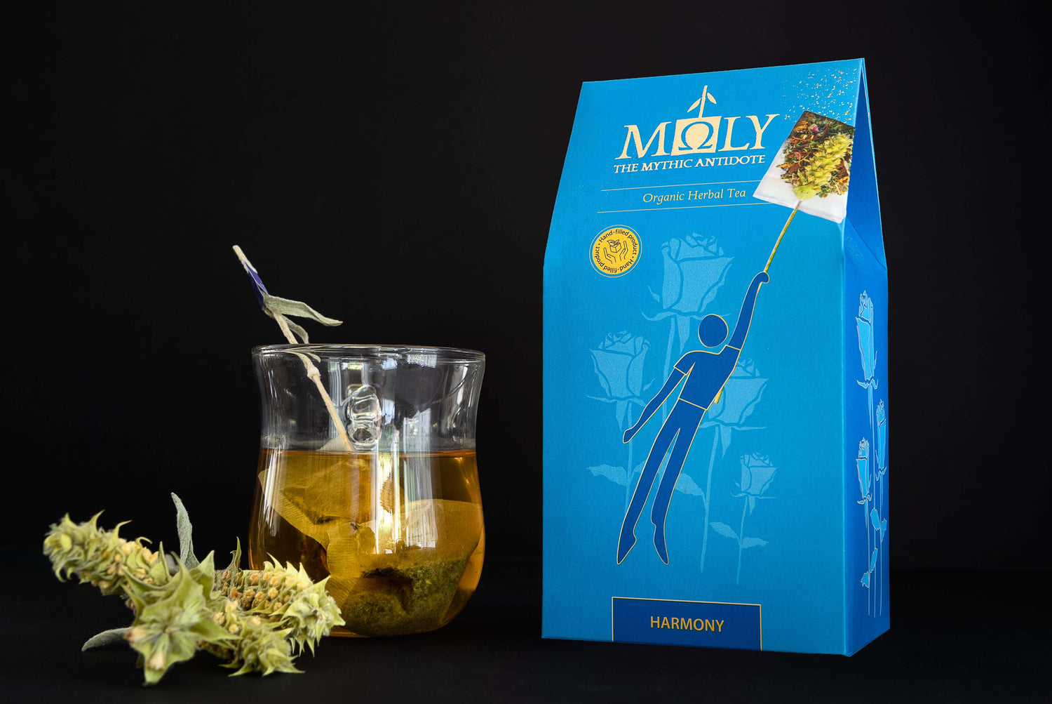 Eine blaue Verpackung des Moly Harmony BIO vor einem schwarzen Hintergrund. Zentral ist ein Bild aufgedruckt, welches einen Teebeutel mit Inhalt zeigt, aus dem der Stengel der Sideritis scardia (griechischer Bergtee) herausragt und auf dem eine Person vom Tee in die Luft gezogen wird. Auf der linken Seite steht ein Tee aus dem der Sideritis -Stengel herausragt. Davor liegen zwei Sideritis-Blüten.