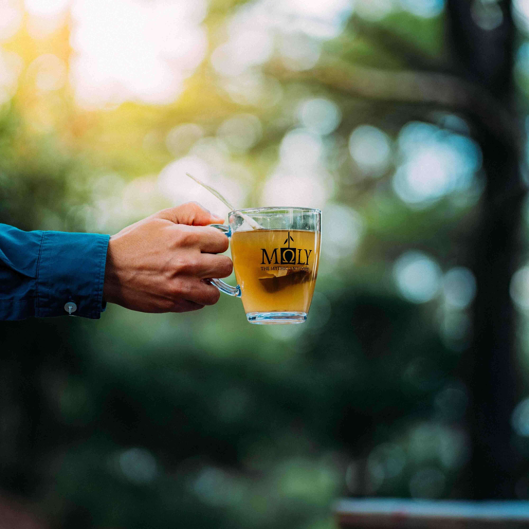 Eine männliche Hand hält ein Glas mit dem MOLY Love. Im Hintergrund sind Bäume zu erkennen.