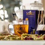 Eine Tasse mit Tee, Bergteeblüten liegen davor. Im Hintergrund die lila Verpackung des MOLY Night Cup