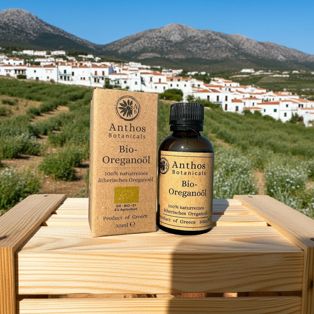 Ein braunes Glasfläschen und Verpackung mit ätherischem Oreganoöl bio  von Anthos Botanicals auf einer Holzkiste vor einem Oreganofeld und einem Dorf.